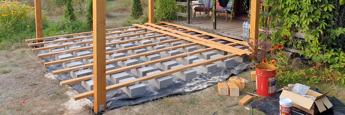 Ruszt legarów tarasowych ułożony na szarych bloczkach betonowych rozstawionych na czarnej agrowłókninie, w tle gotowy taras z drewnianymi słupami