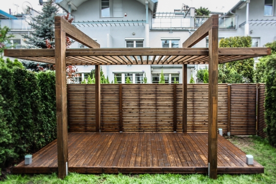 Wolnostojąca pergola z ciemnego drewna na tarasie z desek, w tle ażurowy płot i tuje