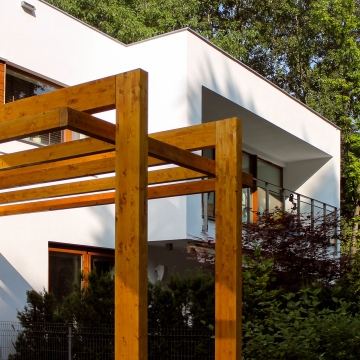 Pergola z drewna w miodowym odcieniu przy białym domu modernistycznym, w tle balustrada tarasu