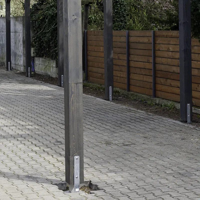 Rząd ciemnych drewnianych słupów osadzonych na stalowych płaskownikach zakotwionych w kostce brukowej, między drewnem a podłożem widoczna szczelina