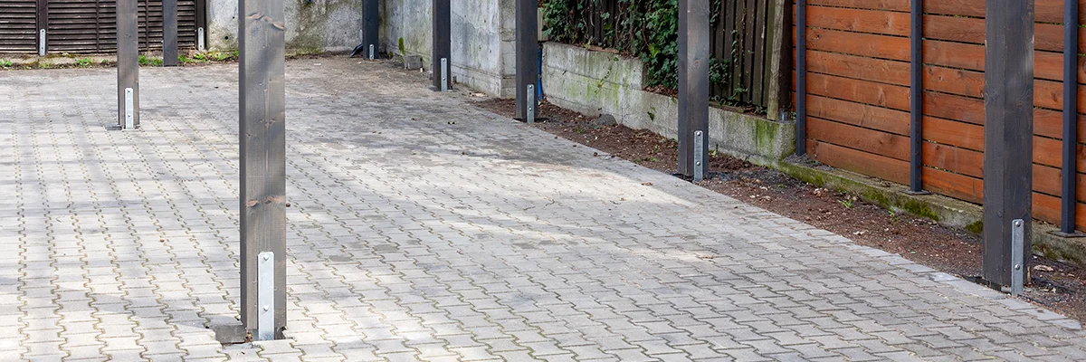 Rząd drewnianych słupów zamocowanych na stalowych kotwach w kostce brukowej, w tle drewniane ogrodzenie