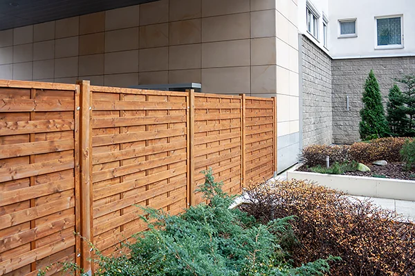 Ogrodzenie z ciemnobrązowych paneli o poziomych deskach wzdłuż podjazdu, po prawej ściana budynku z płyt elewacyjnych