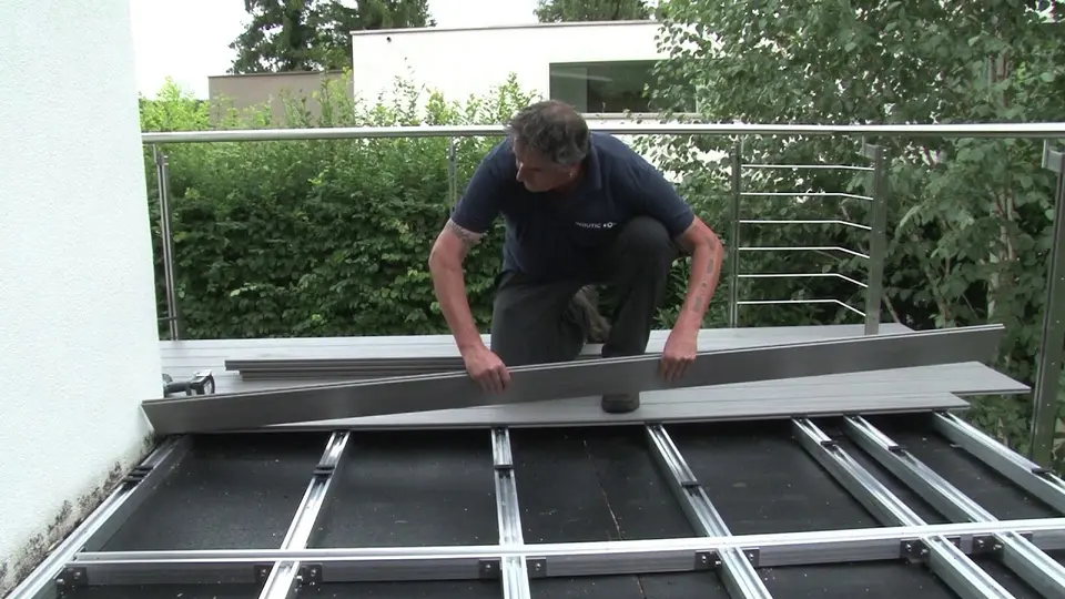 Kadr z filmu: monter układa szarą deskę kompozytową na aluminiowych legarach na balkonie