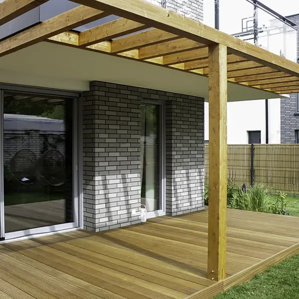 Pergola z jasnych belek klejonych przy przeszklonej ścianie domu, oparta na słupach stojących na drewnianym tarasie