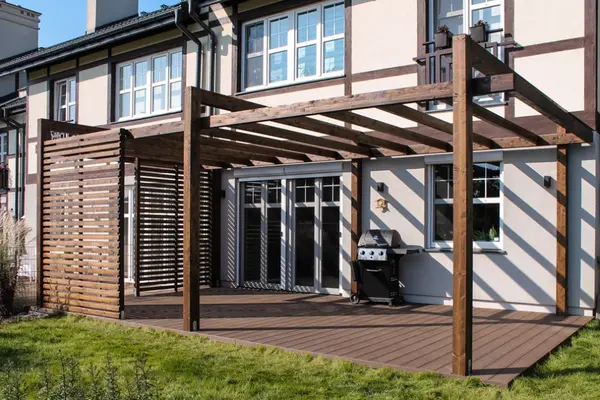 Ciemna pergola z belek klejonych dobudowana do ściany domu, oparta na czterech słupach stojących na brązowym tarasie kompozytowym