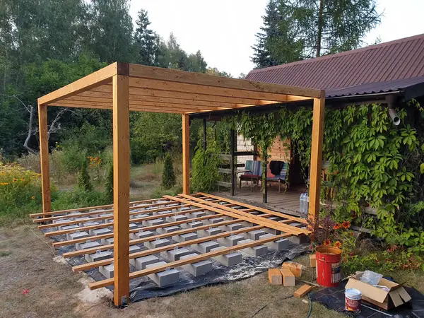 Jasne legary drewniane ułożone na rzędach bloczków betonowych, nad nimi gotowa konstrukcja pergoli z czterech słupów i belek, w tle dom porośnięty winobluszczem