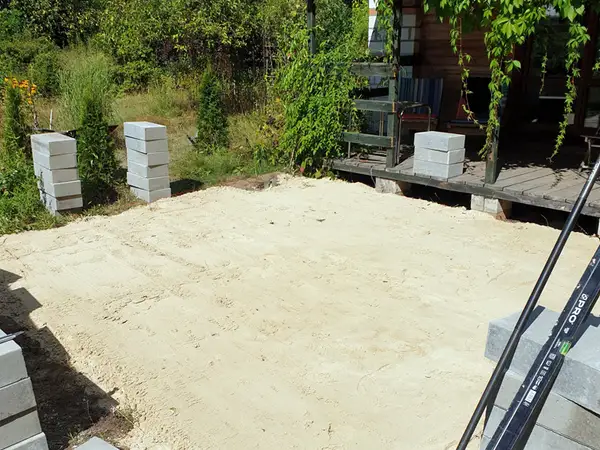 Wyrównana warstwa jasnego piasku przed drewnianym domem z werandą, wokół niej stosy szarych bloczków betonowych, na pierwszym planie długa poziomica