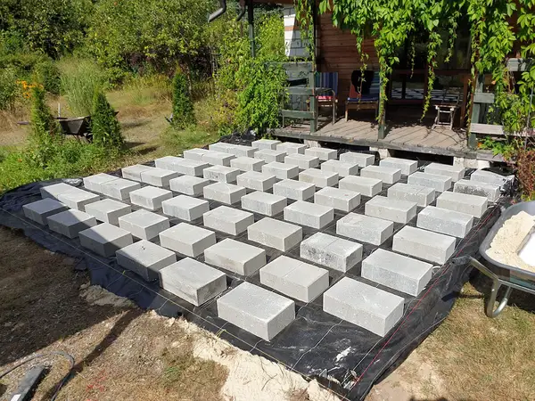 Równe rzędy szarych bloczków betonowych rozstawionych na czarnej włókninie, między nimi naciągnięte czerwone sznurki, obok taczka z piaskiem