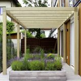 Pergola z jasnego drewna nad podestem między budynkami, pod nią donica z trawami ozdobnymi